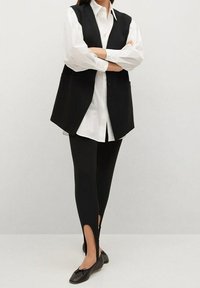 Schwarze ärmellose Weste über einem weißen Hemd mit Knöpfen, kombiniert mit schwarzen Leggings mit seitlichen Schlitzen und schwarzen flachen Schuhen.