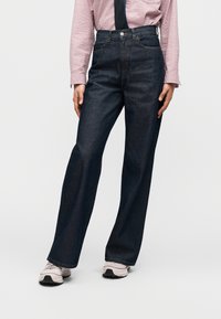 Τζιν παντελόνι με φαρδιά μπατζάκια σε σκούρο τζιν ύφασμα, με κλασικό σχεδιασμό πέντε τσεπών, συνοδευόμενο από διακριτική μπλε απόχρωση και ομαλή υφή.