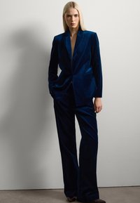 Blauveloursblazer und passende Hose mit taillierter Passform. Der Blazer hat Spitzrevers und seitliche Taschen, die das strukturierte Design unterstreichen.