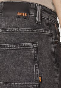 Närbild av mörk denimjeans med en svart etikett där det står "BOSS" i orange, samt en synlig ficka p å baksidan.