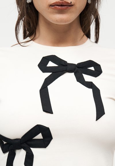 Γυναίκα που φοράει λευκό τοπ με δύο μαύρα υφασμάτινα φιογκάκια, ένα κοντά στη λαιμόκοψη και ένα κοντά στο στήθος, με ορατά σκουλαρίκια και κραγιόν.