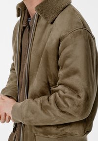 Hnědá bomber bunda z semiše s měkkým, texturovaným povrchem, žebrovanými manžetami a kontrastním límcem z ovčí vlny. Přední zapínání na zip.