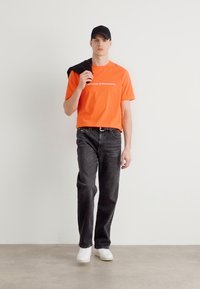 Oranžové grafické tričko s bílým textem, černé džínové kalhoty, černá čepice a bílé tenisky. Model stojí na neutrálním pozadí.