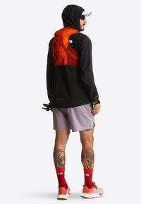 Czarna wodoodporna kurtka, pomarańczowa kamizelka nawadniająca, jasnofioletowe spodenki, czerwone skarpety i buty. Widoczne tatuaże na nogach. Dobra widoczność i funkcjonalny design.