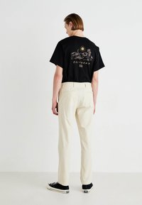 Tricou negru din bumbac cu un print grafic care prezintă munți și cuvântul "California" în aur; asortat cu pantaloni bej și adidași negri.