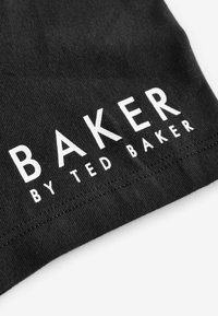 Fekete szövetcímke, amelyen fehér szöveg olvasható: "BAKER" és "by TED BAKER". A címke texturált felületű és varrás díszíti az éleit.