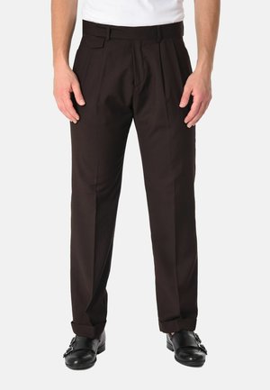Uomo con pantaloni neri su misura, scarpe eleganti in pelle nera con fibbie e una camicia bianca su uno sfondo semplice.