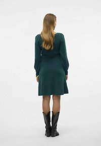 Knälång klänning med långa ärmar i mörkt teal-stickat, med en figursydd byst och utsvängd kjol. Bärs med svarta lädercowboystövlar.