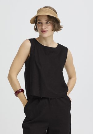 Frau trägt ein schwarzes ärmelloses Oberteil und Hosen mit einer beigefarbenen Schirmmütze und einem dunkelroten Armband, posiert mit den Händen in den Taschen.