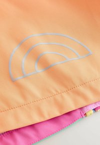 Približan pogled na breskvinasto tkanino z reflektivnim polkrožnim vzorcem nad všitim robom ter plasti roza in zelene tkanine spodaj.