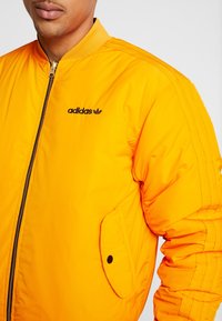 Klart orange bomberjacka med dragkedja, ribbat krage, logotyp på bröstet, sidoficka med klaff och synliga sömdetaljer.