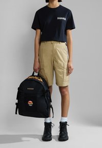 Zaino nero con tasca in rete e toppa multicolore, abbinato a una maglietta blu navy e pantaloni cargo beige, indossato con stivali neri stringati.