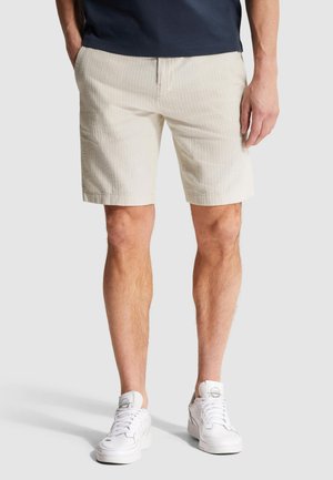 Mand iført beige shorts, mørkeblå skjorte og hvide sneakers står med hænderne i lommerne mod en lys, ensfarvet baggrund. Kun underkroppen er synlig.