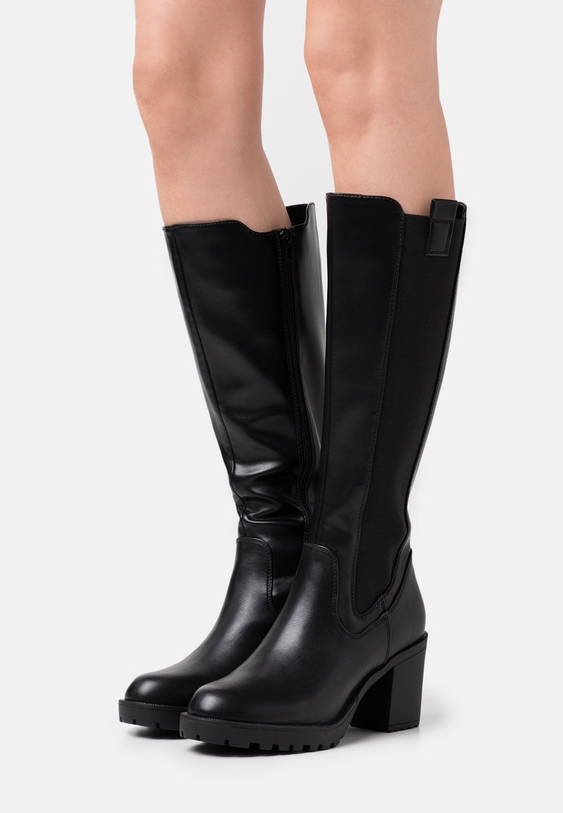 Jambes portant des bottes en cuir noir montantes jusqu'aux genoux, à talons blocs et fermeture éclair latérale, présentées sur un fond clair uni.