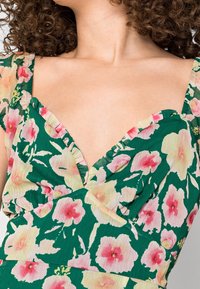 Blommig klänning med grön bakgrund och rosa och gula blommor. V-ringad design och volangärmar i texturerad tyg.