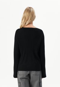 Blonde woman facing backward wearing a black ribbed sweater and gray cargo pants against a white background.