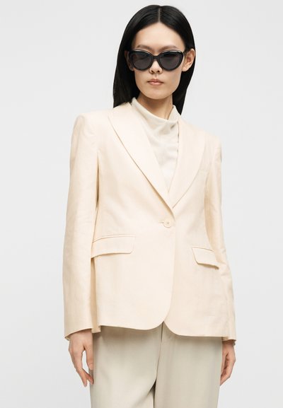 Kvinde med glat sort hår bærer sorte solbriller, en off-white blazer og matchende højtaljede bukser mod en ensartet baggrund.