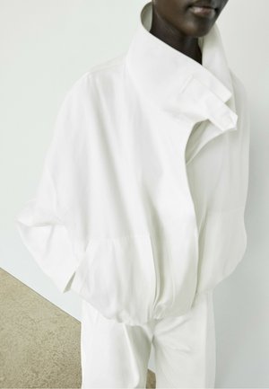 Personne à la peau foncée portant une veste blanche ample avec un col montant et un pantalon blanc assorti, debout devant un fond clair uni.