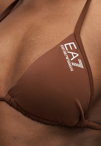 Καφέ τρίγωνο πάνω μέρος μπικίνι με λευκό λογότυπο EA7 Emporio Armani, φορεμένο πάνω σε μαύρο δέρμα.