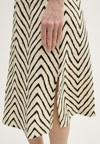 Ein knielanges Kleid aus leichtem Material, das einen cremigen Grundton mit auffälligen schwarzen Zickzackmustern und einem Seiten Schlitz für mehr Bewegungsfreiheit aufweist.