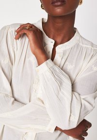 Ingrandimento di una persona che indossa una blusa bianca a maniche lunghe con un delicato ricamo floreale, orecchini d'oro e anelli su entrambe le mani.