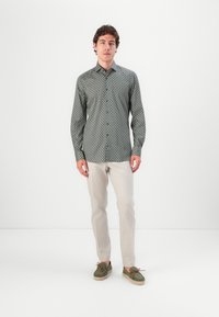 Herrenhemd mit Muster, das dunkle grüne geometrische Designs auf einem hellen Hintergrund zeigt, kombiniert mit hellbeigen Hosen und grünen Schuhen.