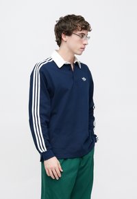 Tengerészkék hosszú ujjú galléros póló fehér gallérral, fehér függőleges csíkokkal az ujjak mentén és egy kis logóval a mellkason.