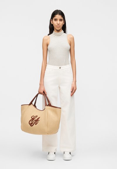 Νεαρή γυναίκα που φοράει ριγέ ζιβάγκο χωρίς μανίκια και λευκό παντελόνι, κρατώντας μια μεγάλη μπεζ τσάντα ώμου με καφέ δερμάτινα χερούλια.