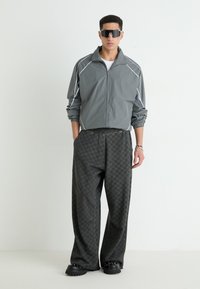Veste coupe-vent grise avec des accents blancs, assortie à un pantalon large à carreaux sombres et des chaussures noires épaisses. Les accessoires incluent des lunettes de soleil angulaires.