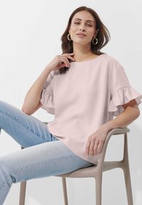 Helles rosa Blusenshirt mit rüschierten Ärmeln, Rundhalsausschnitt, glatter Textur. Kombiniert mit hellblauen Jeans. Modell sitzt auf einem beigen Stuhl.