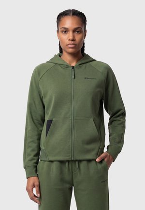 Donna con capelli intrecciati che indossa una felpa verde oliva con zip e pantaloni della tuta abbinati, in piedi con una mano in tasca davanti a uno sfondo semplice.