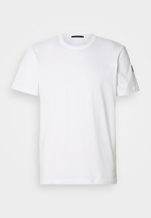 T-shirt in cotone bianco con colletto rotondo e maniche corte; presenta un logo discreto sulla manica sinistra. Texture liscia e vestibilità regolare.