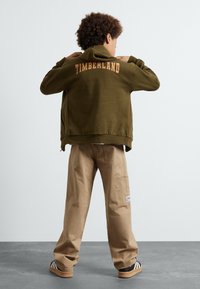 Olivově zelená mikina s kapucí se znakem "TIMBERLAND" na zadní straně, v kombinaci s béžovými volnými cargo kalhotami a černými proužkovanými pantoflemi.