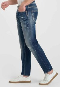 Jeans de mezclilla en azul oscuro con un lavado desvanecido y efectos de arrugas, que cuentan con cinco bolsillos y detalles de costura en color naranja. Combinados con zapatillas blancas.