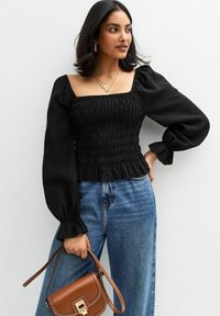 Top nero con maniche a sbuffo, texture arricciata e scollatura quadrata, abbinato a jeans chiari a gamba larga e una piccola borsa a tracolla marrone.
