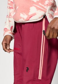 Vinrøde vandtætte bukser med rosa lynlåsdetalje og sort logo. Har sidelommer og et elegant, tilspidset design.