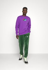 Lilla sweatshirt med en Nike-logo patch, parret med grønne velour sweatpants med hvide sidelister og et lille logo. Sorte høj-top sneakers.