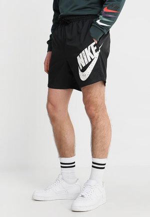 Mężczyzna w czarnych szortach Nike z dużym białym logo, białych skarpetkach w paski i białych butach sportowych Nike, stojący z ręką w kieszeni.