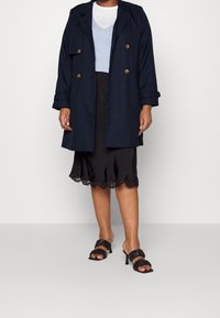 Cappotto trench navy con bottoni doppi, indossato sopra un top blu chiaro e una gonna nera di satin con orlo in pizzo. Sandali neri con tacco.