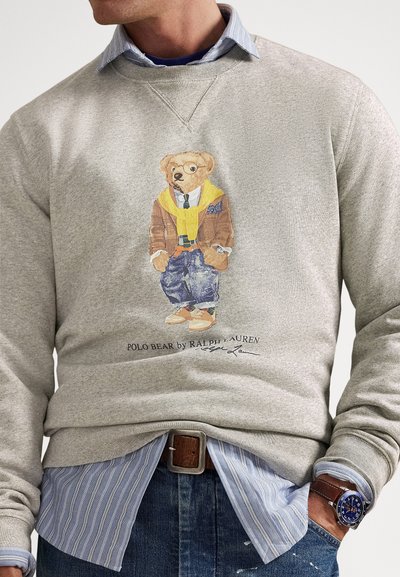 Γκρι φούτερ με αρκούδα που φοράει ταμπά σακάκι, κίτρινο πουλόβερ και τζιν. Σημαντικές λεπτομέρειες λογότυπου και εικαστικών στο κάτω μέρος.
