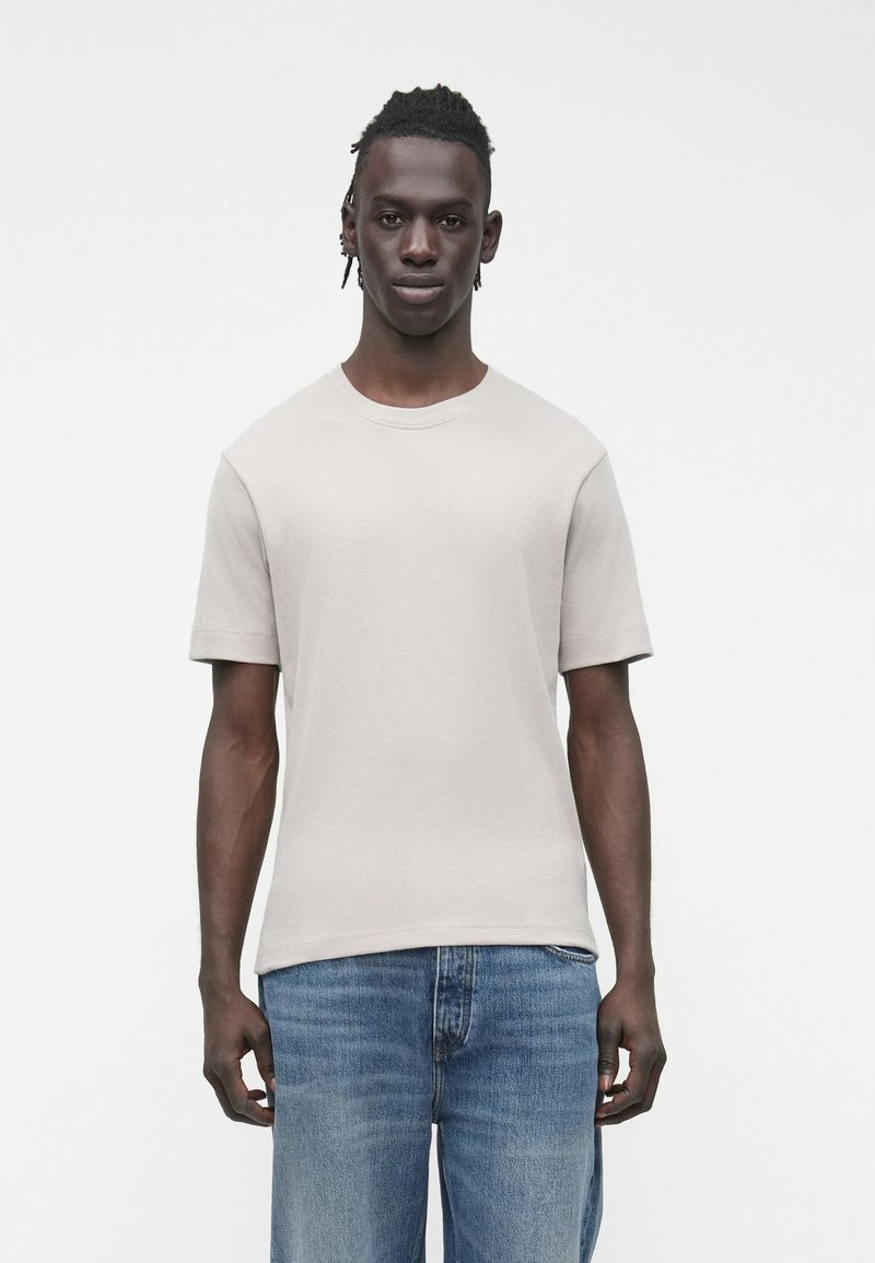 Tricou cu mânecă scurtă, culoare bej deschis, cu o textură striată și gât rotund, asortat cu blugi din denim albastru mediu. Estetică simplă și curată.