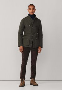 Jakna od naquiltanog maslinastozelenog materijala s okovratnikom, prednjim džepovima i sivoj akcentima. Uparen s smešnim hlačama i smešnim čizmama na vezivanje.