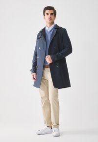 Hombre vestido con un abrigo azul marino de doble botonadura sobre un suéter y una camisa azul, pantalones beiges y zapatillas blancas, de pie frente a un fondo claro y liso.