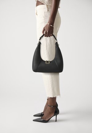 Personne tenant un sac à main en cuir noir texturé avec un logo doré, portant un pantalon blanc cropé et des talons hauts noirs pointus avec des lanières à la cheville.