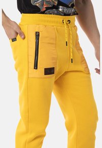Gelbe Jogginghose aus glattem Material, mit elastischem Bund, gelbem Kordelzug und seitlichen Reißverschlusstaschen mit schwarzen Akzenten.