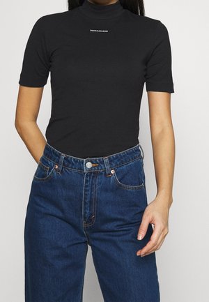 Maglietta nera a maniche corte con collo alto e piccolo logo "Calvin Klein Jeans", abbinata a jeans blu scuro larghi e a vita alta.