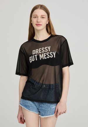 T-shirt in rete nera con la scritta "DRESSY BUT MESSY", caratterizzato da maniche corte e un orlo curvo, indossato sopra un design di pantaloncini in denim stampati.