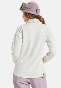 Biała bluzka z długim rękawem z teksturowanym wzorem, wysokim kołnierzem i logo na rękawie. W zestawie z jasnofioletowymi spodniami.