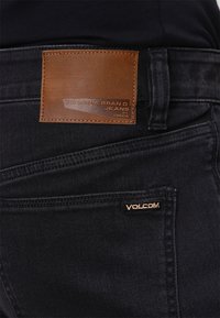 Pantalones vaqueros negros con un bolsillo trasero que tiene un parche de cuero marrón etiquetado como "VOLCOM BRAND JEANS" y un pequeño logotipo bordado de "VOLCOM" en el borde del bolsillo.