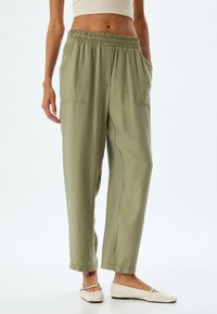 Lichtgroene, losse broek met een elastische tailleband, zijzakken en een gladde textuur, gedragen met platte, beige schoenen.
